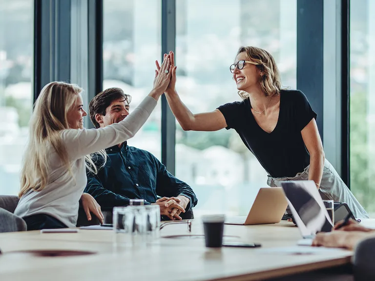 Trois personnes dans une salle de réunion lumineuse se félicitent par un high five, devant des ordinateurs portables et des tasses sur la table.