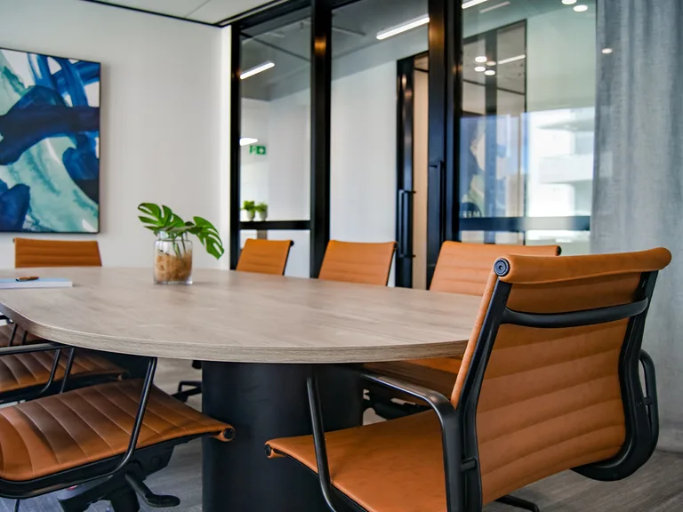 A modern office conference room with a wooden table, orange chairs, a potted plant, and an abstract painting on a white wall. Large glass windows and door surround the space.