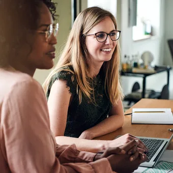 Two people sit at a wooden table in an office, one wearing a pink top and the other a green lace top with glasses, smiling at a laptop.