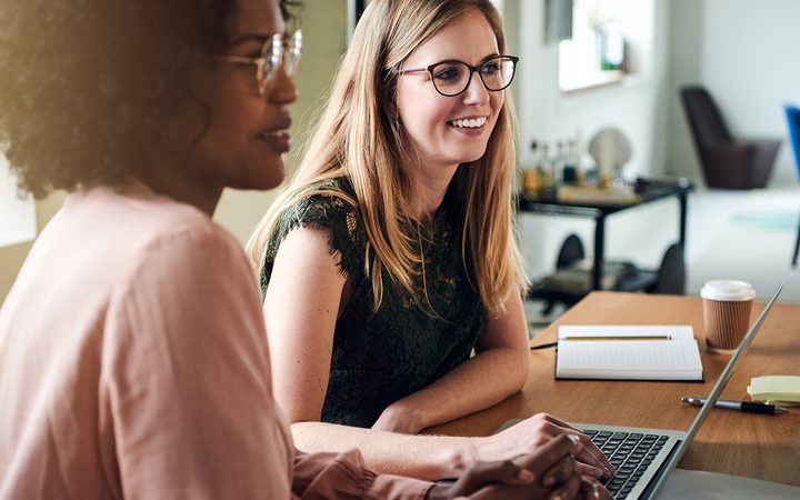 Two people sit at a wooden table in a bright office, smiling at a laptop; one wears glasses and a green lace top, the other has curly hair.