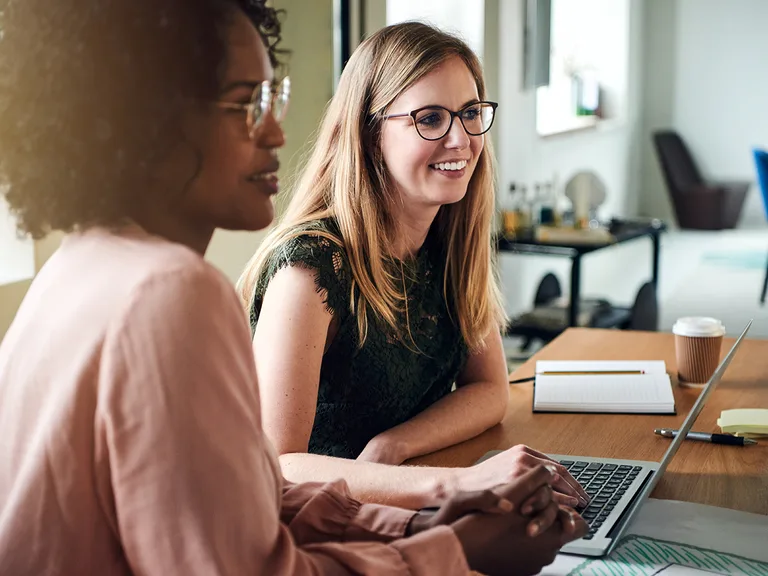 Deux personnes autour d'une table; l'une porte des lunettes et un haut vert en dentelle, l'autre en rose pâle, devant un ordinateur portable.