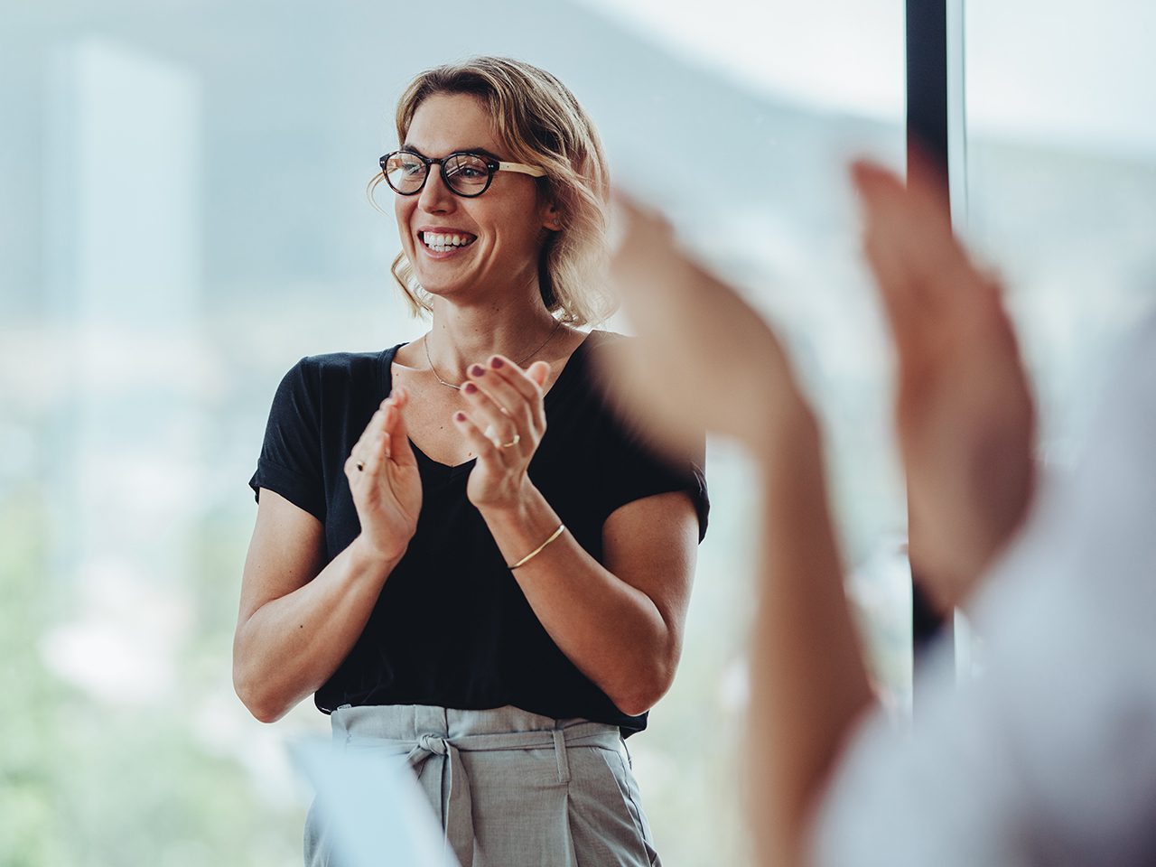 Smiling person with short light hair and glasses claps in a bright room, with blurred hands and a window in the background.