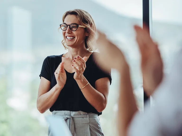 Smiling person with short light hair and glasses claps in a bright room, with blurred hands and a window in the background.