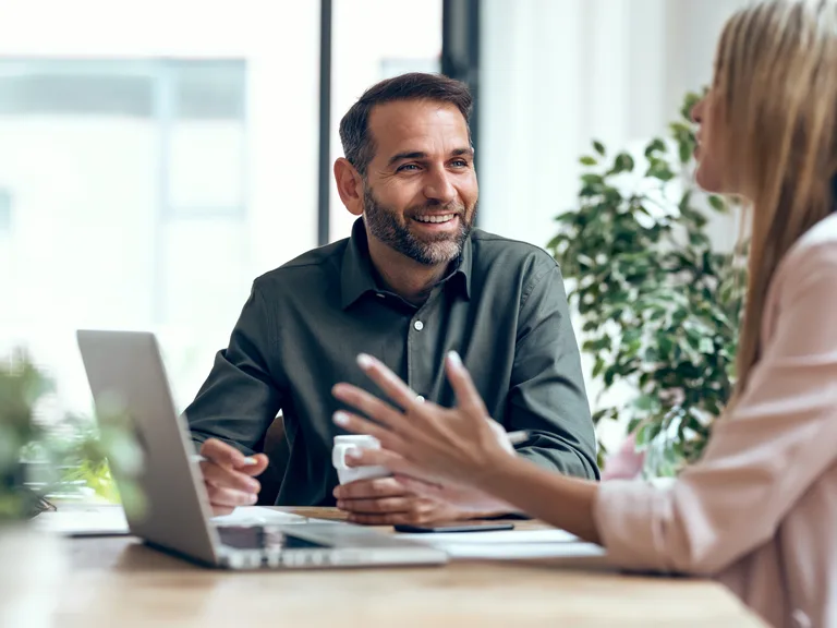 Two people sit at a bright table with a laptop, smiling and talking. One has short dark hair and a beard; the other has long hair in a pale pink top.