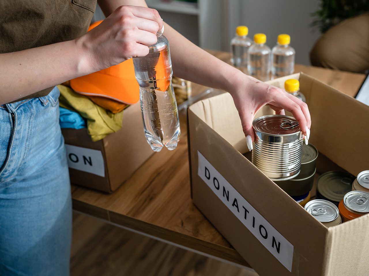 A person is packing canned food and bottled water into donation boxes on a wooden table.