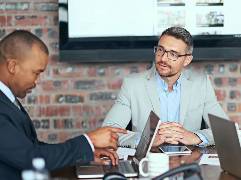 Two professionals in a meeting at a glass table with laptops; one points at a laptop, the other sits with hands folded, beside a brick wall and screen.