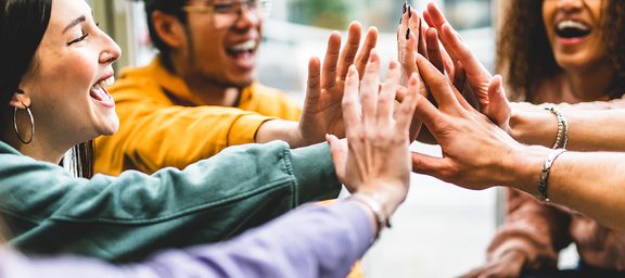 Diverse group of friends high-fiving with hands joined in the center, smiling and laughing indoors.
