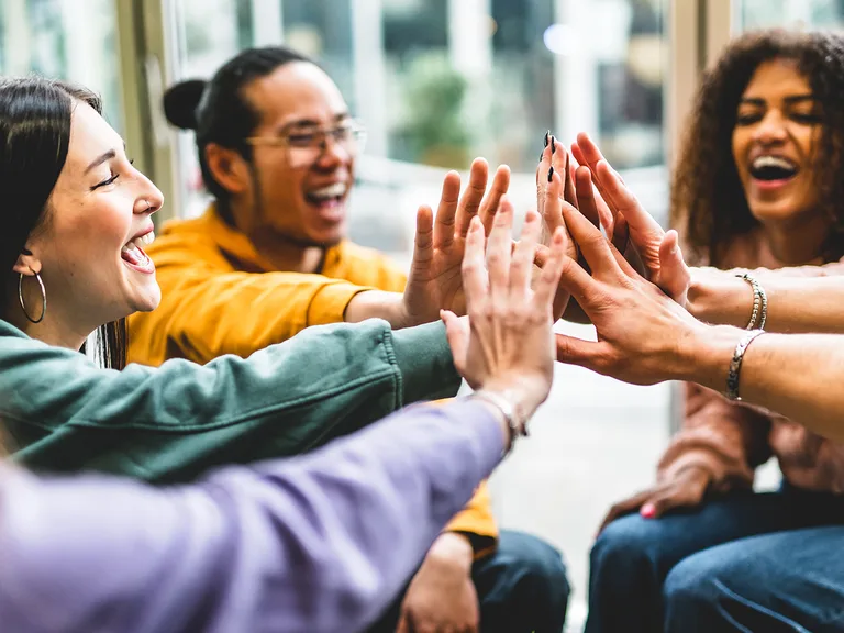 Group of diverse friends giving high-fives, smiling and laughing indoors by a window.