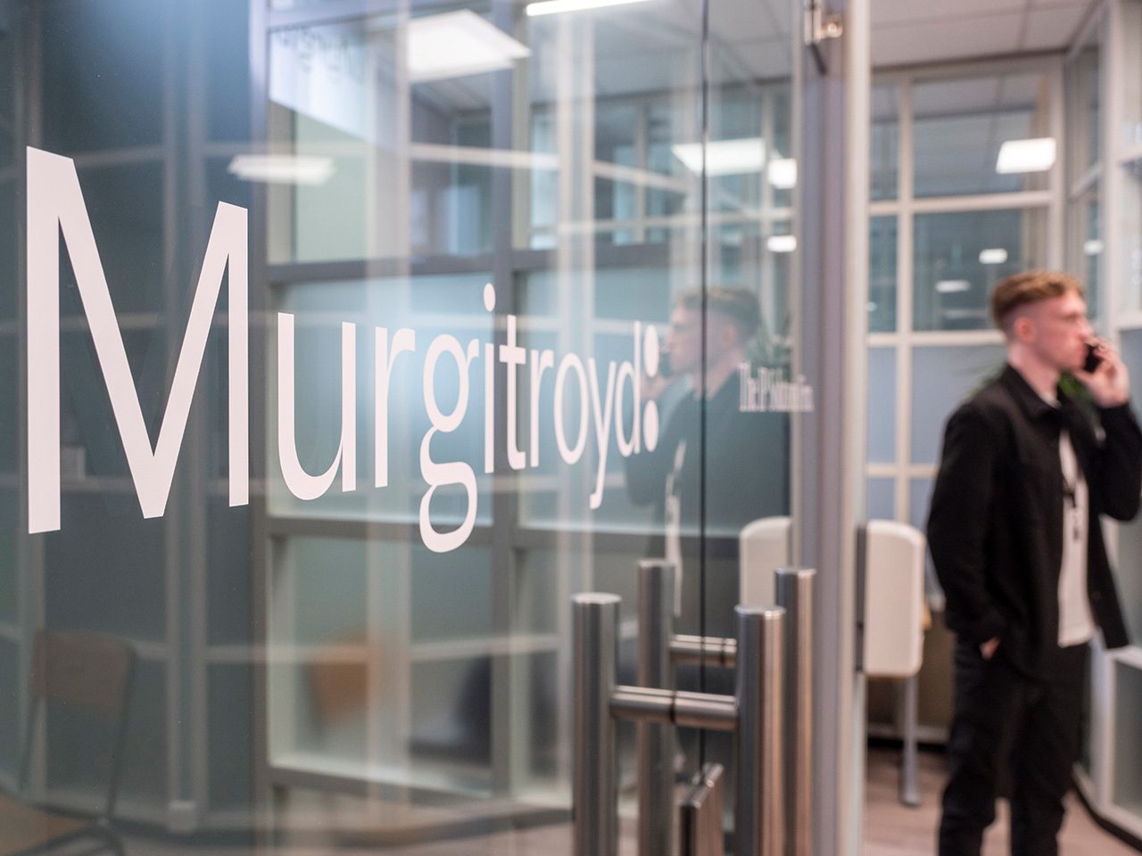 Office lobby with a glass wall bearing large white 'Murgitroyd' lettering; a person in dark clothing stands on the right, talking on a mobile phone.