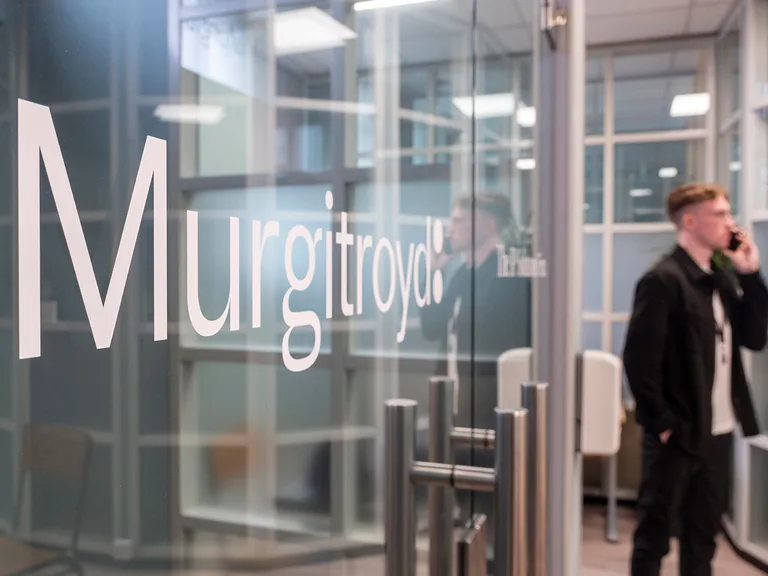 Office lobby with a glass wall bearing large white 'Murgitroyd' lettering; a person in dark clothing stands on the right, talking on a mobile phone.
