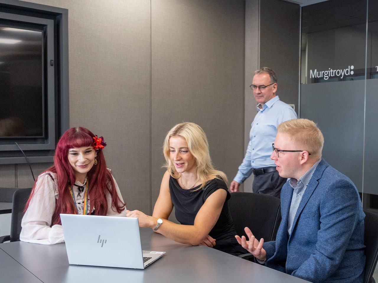 Four colleagues in a meeting room sit around a silver HP laptop; two focus on the screen while another gestures, and a fourth stands by a glass wall.