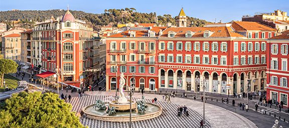 A lively city square with red and pink historic buildings, a central fountain with statues, and people walking on a patterned pavement in bright sunlight.