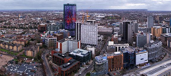 Aerial view of a city skyline with various modern high-rise buildings and a digital billboard, extending into a densely built urban landscape.