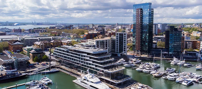 A cityscape with modern buildings, a marina filled with boats, and a body of water, under a partly cloudy sky.