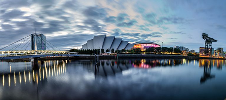 A city skyline at dusk with modern buildings, calm water reflecting the lights, and a partly cloudy sky overhead.