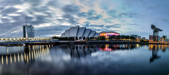 Sky and city skyline reflected in calm water, with a bridge on the left, a modern building in the center, and a stadium and cranes on the right.