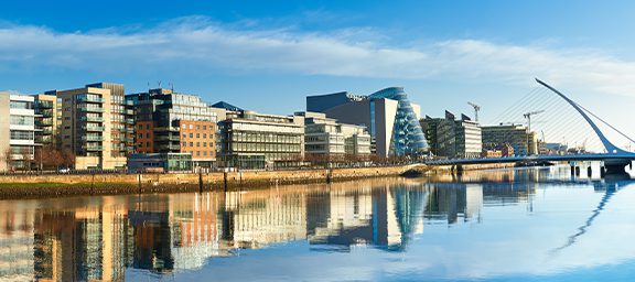 City skyline with modern buildings reflected in a calm river under a blue sky. A bridge with a curved design is visible on the right side.