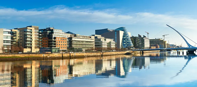 Modern cityscape with tall buildings along a river, reflected in the water, under a partly cloudy sky.