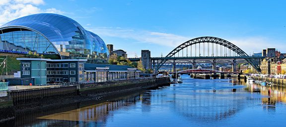 City skyline featuring a modern glass building, historic bridge, and calm water reflecting the structures under a clear blue sky.