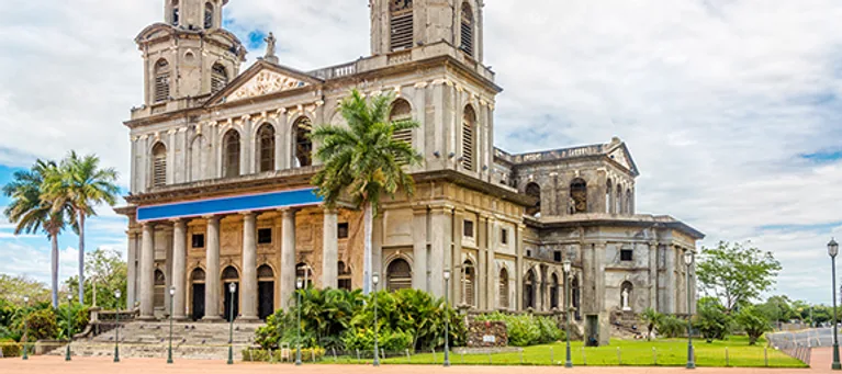 A historic stone church with twin bell towers, surrounded by palm trees and greenery, under a partly cloudy sky.