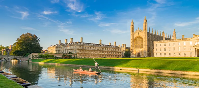 A person kayaking on the river beside historic university buildings, including a large Gothic-style chapel, under a bright blue sky.