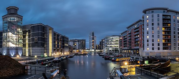 A city skyline along a river at dusk, featuring modern buildings with illuminated windows and boats docked along the water.