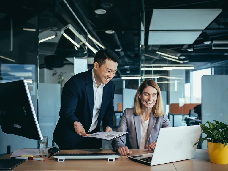 Deux collègues dans un bureau moderne: une personne debout montre des documents à l'autre, près d’un ordinateur portable, sourire partagé.