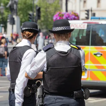 Portrait of a person with light brown hair and blue eyes, wearing a blue sleeveless top with two black buttons, smiling against a grey backdrop.