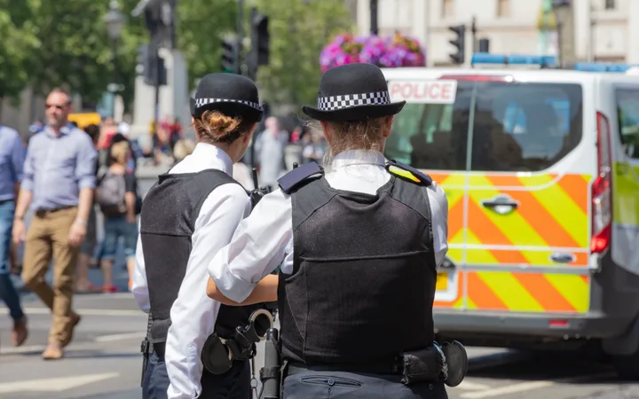 Two police officers with black hats and vests stand on a busy city street, with a police vehicle and pedestrians in the background.