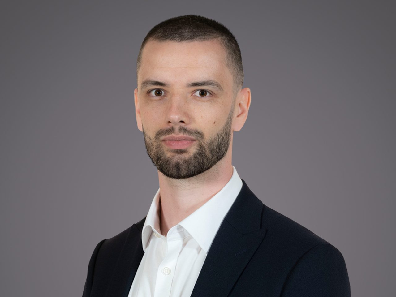 Portrait of a person with short dark hair and a beard, wearing a white shirt and dark blazer, looking at the camera against a gray studio backdrop.