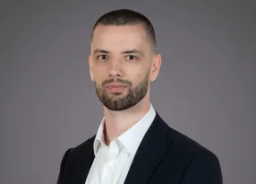 Portrait of a person with short dark hair and a trimmed beard, wearing a white shirt and dark blazer, posed against a grey studio background.