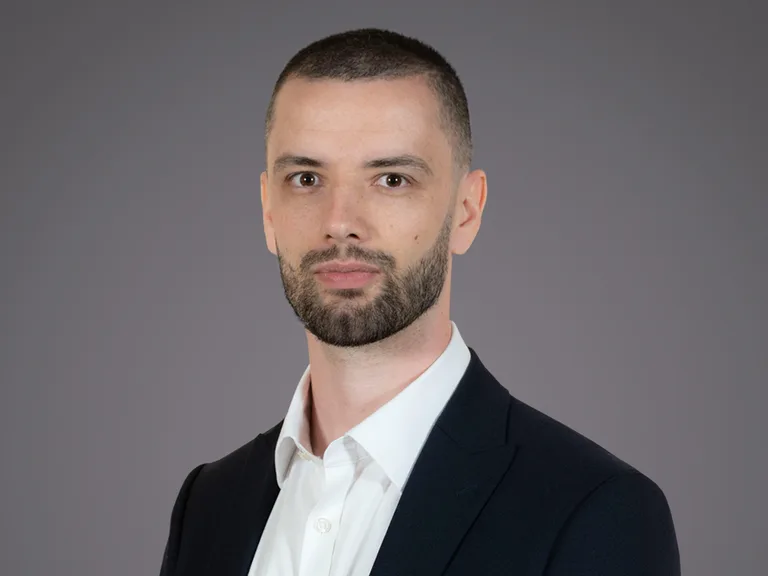Homme d'affaires avec une barbe, portant un costume noir et une chemise blanche, posé contre un fond gris, regardant l'objectif avec sérieux.