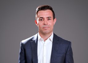 Portrait of a person in a navy blazer and white shirt, with short dark hair, against a grey gradient studio background.