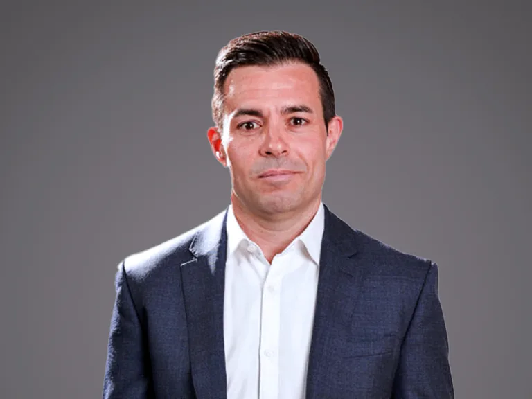 Portrait of a person in a navy blazer and white shirt, with short dark hair, against a grey gradient studio background.