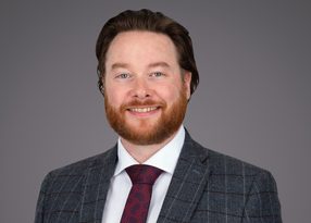 Portrait of a smiling person with red hair and beard, wearing a gray checkered blazer, white shirt, and maroon tie, against a neutral gray background.