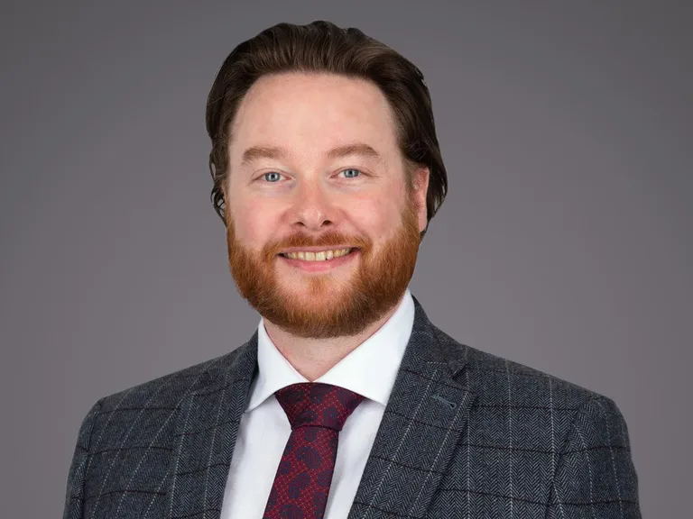 Smiling person with a red beard and blue eyes, wearing a grey checked blazer, white shirt and burgundy tie, against a neutral grey background.