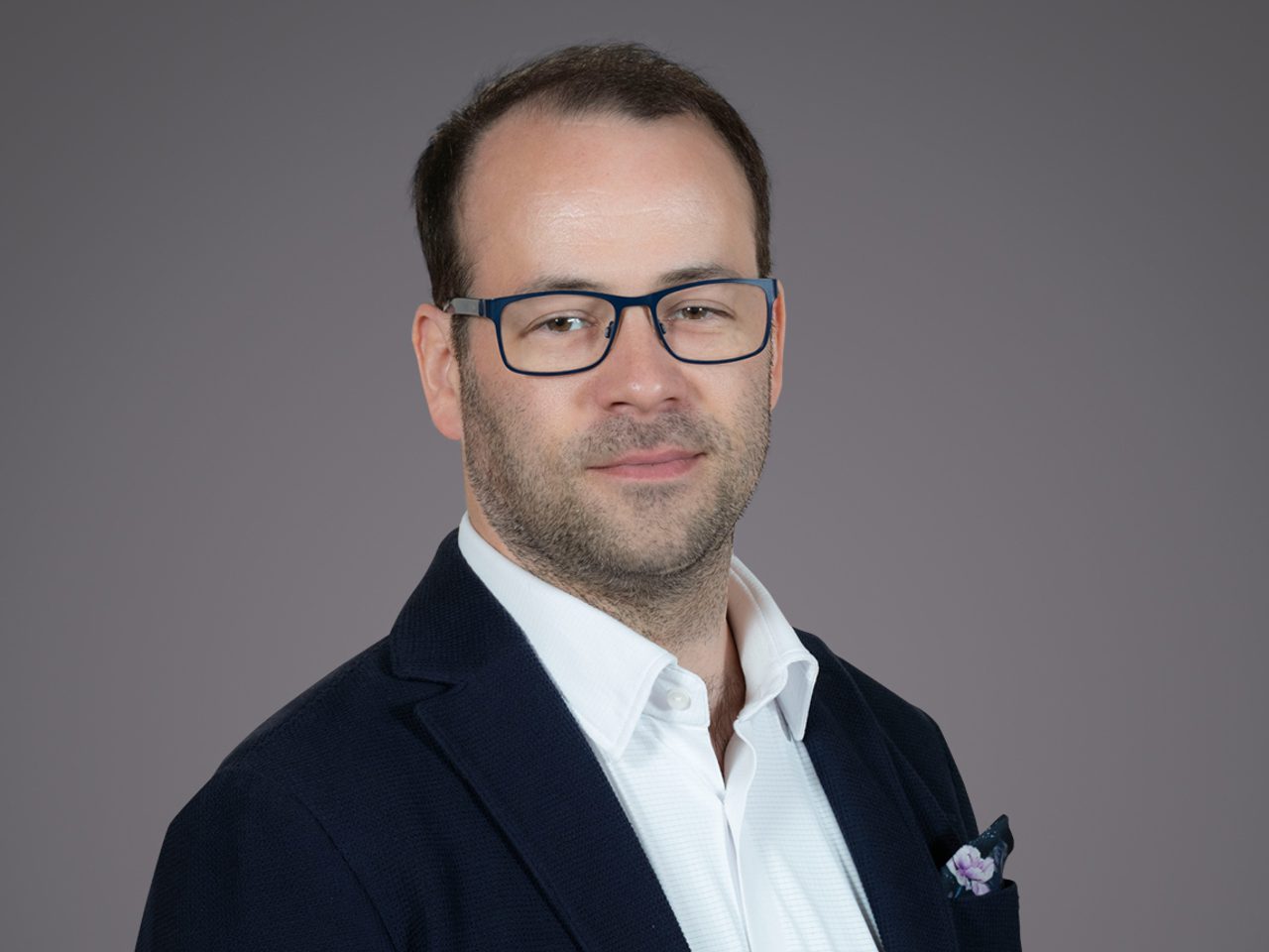 Portrait of a person with short dark hair and blue-framed glasses, wearing a navy blazer, white shirt, and a lilac pocket square, posed against a gray background.