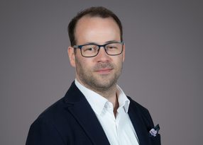 Portrait of a person with short dark hair and blue-framed glasses, wearing a navy blazer, white shirt, and a lilac pocket square, posed against a gray background.