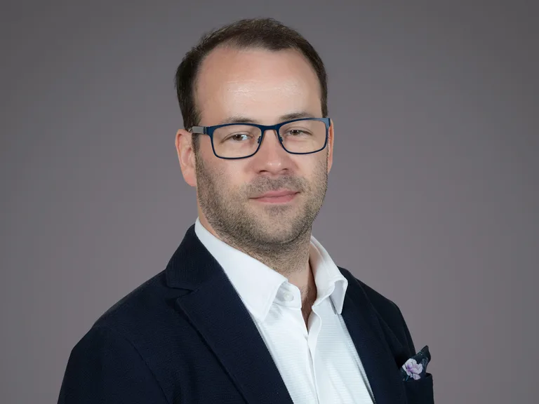 Portrait of a person with short dark hair and blue-framed glasses, wearing a navy blazer, white shirt, and a lilac pocket square, posed against a gray background.