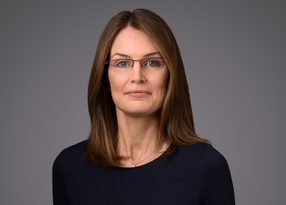 Portrait of a person with shoulder-length brown hair and glasses, wearing a dark navy top, against a grey studio backdrop.