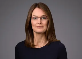 Portrait of a person with shoulder-length brown hair and glasses, wearing a dark navy top, against a grey studio backdrop.