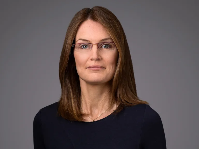 Portrait of a person with mid-length light brown hair and red-framed glasses, wearing a dark navy top, neutral expression against a gray backdrop.