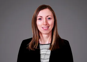 Portrait of a person with shoulder-length light brown hair, wearing a black blazer over a white and black patterned top, smiling.