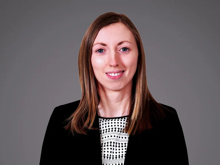 Portrait of a person with shoulder-length light brown hair, wearing a black blazer over a white and black patterned top, smiling.