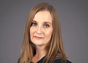 Portrait of a person with straight light brown hair, wearing a dark top, gazing at the camera with a neutral expression against a grey background.