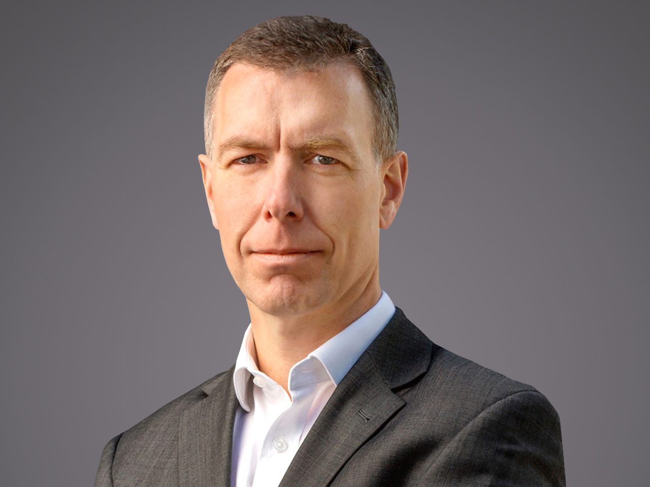 Portrait of a person in a dark suit and white shirt, facing the camera with a neutral expression, against a grey gradient background.