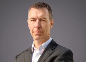 Portrait of a person in a dark suit and white shirt, facing the camera with a neutral expression, against a grey gradient background.