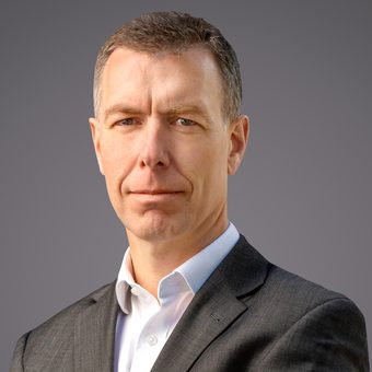 Portrait of a person in a dark suit and white shirt with a neutral expression, short hair, against a gray gradient background.