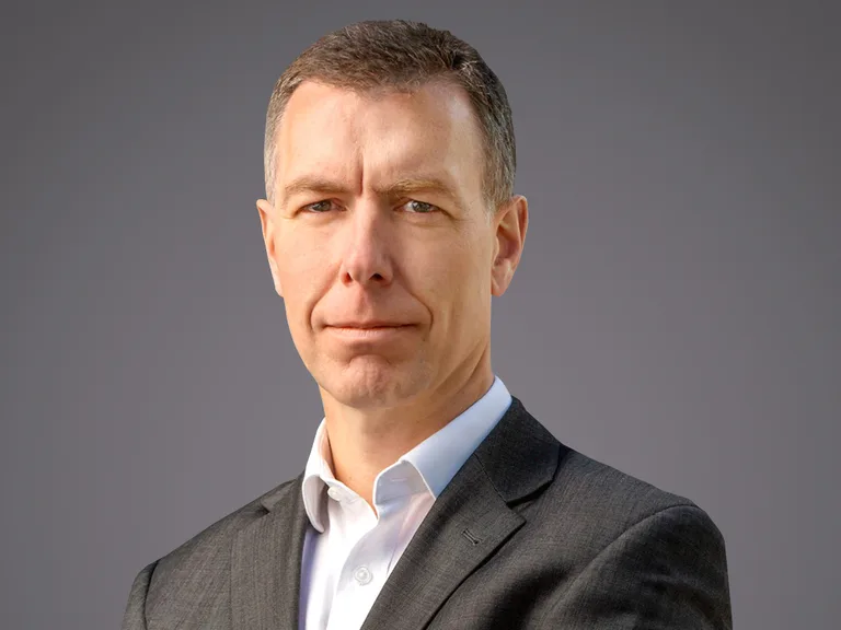 Portrait of a person in a dark suit and white shirt, facing the camera with a neutral expression, against a grey gradient background.