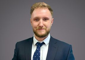 Professional headshot of a person with short blond hair and beard, wearing a navy suit, white shirt, and blue patterned tie, against a gray gradient background.